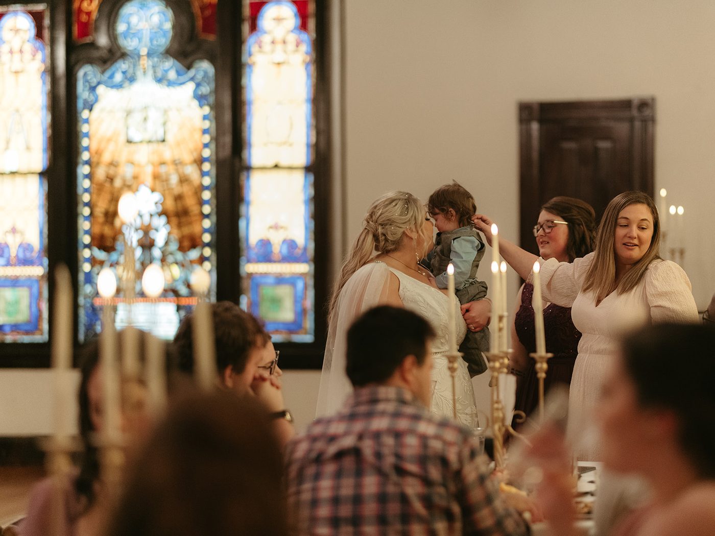 A bride holding a small child among guests at a candlelit table, the Sanctuary's stained glass lit behind them