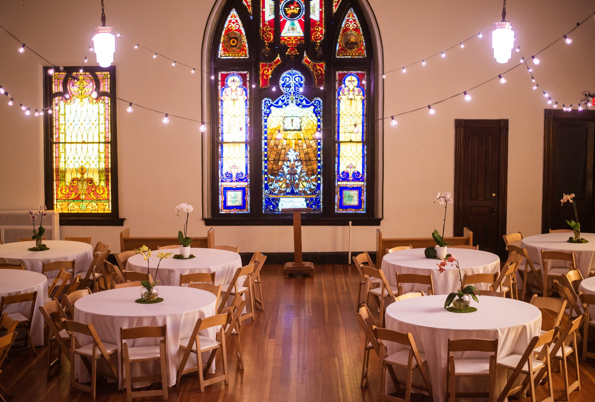 The Sanctuary laid out for a wedding reception — round tables under strings of café lights, facing the great stained-glass window